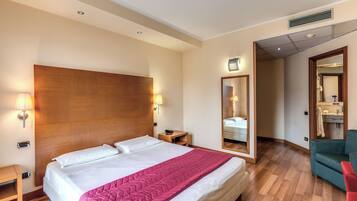 A hotel room with a large bed, wooden headboard, bedside lamps, a mirror, a red chair, and a wooden floor.