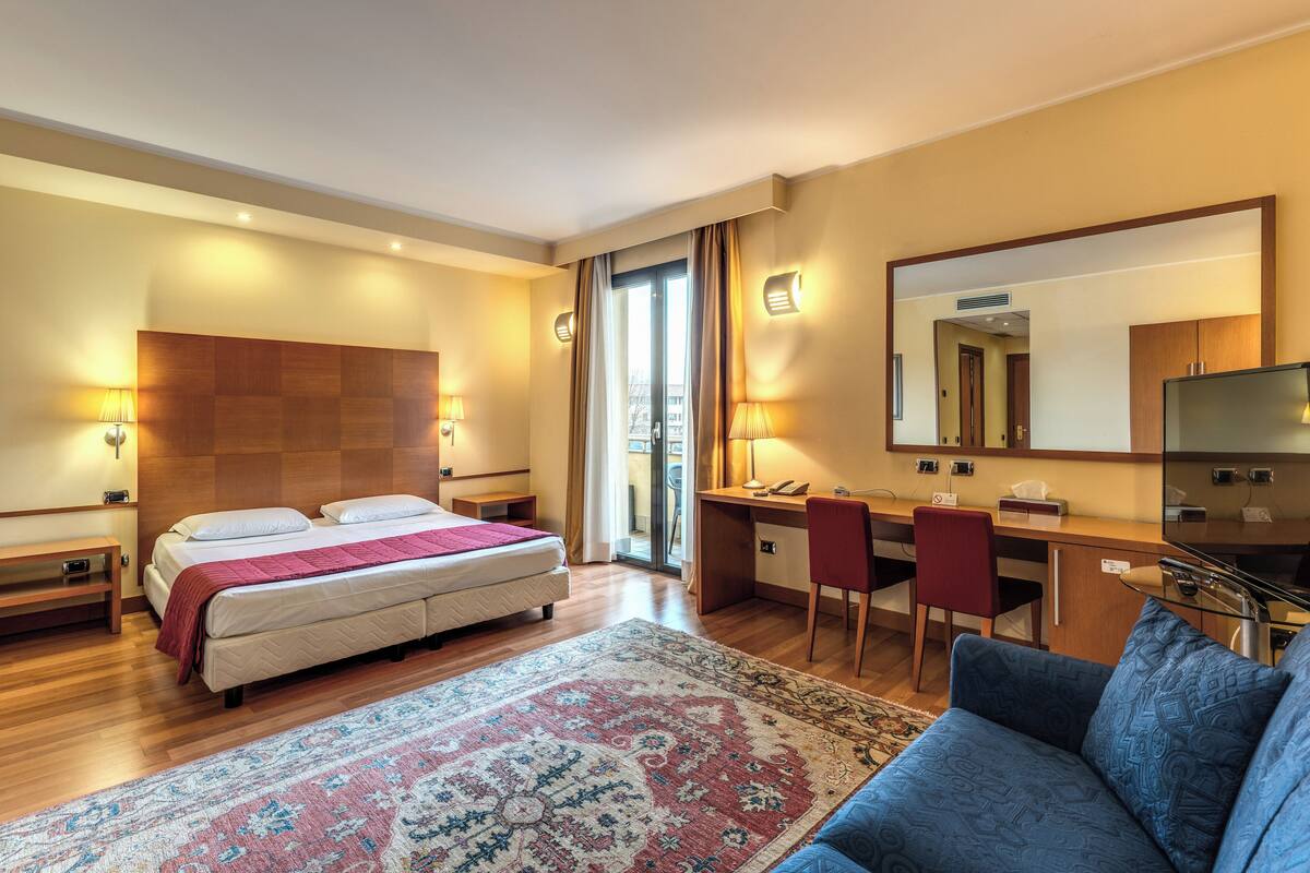 A hotel room with a bed, desk, chair, and a large mirror.