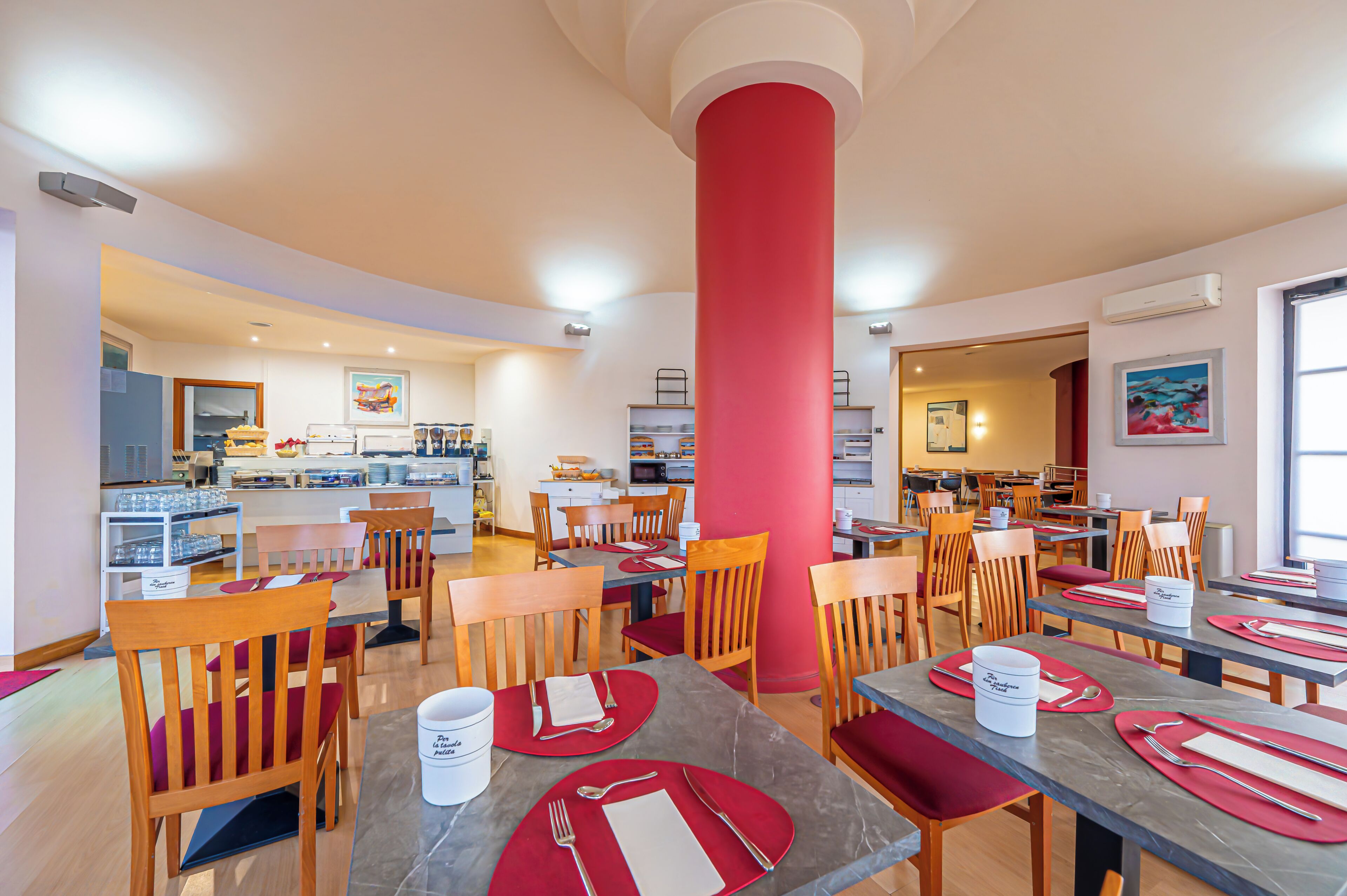A hotel dining area with multiple tables and chairs, a central red pillar, and a kitchen in the background.