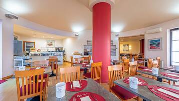 A hotel dining area with multiple tables and chairs, a central red pillar, and a kitchen in the background.