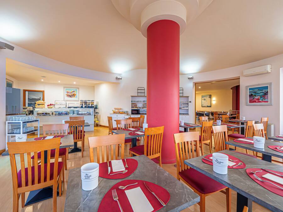 Un comedor de hotel con varias mesas y sillas, un pilar rojo central y una cocina al fondo.