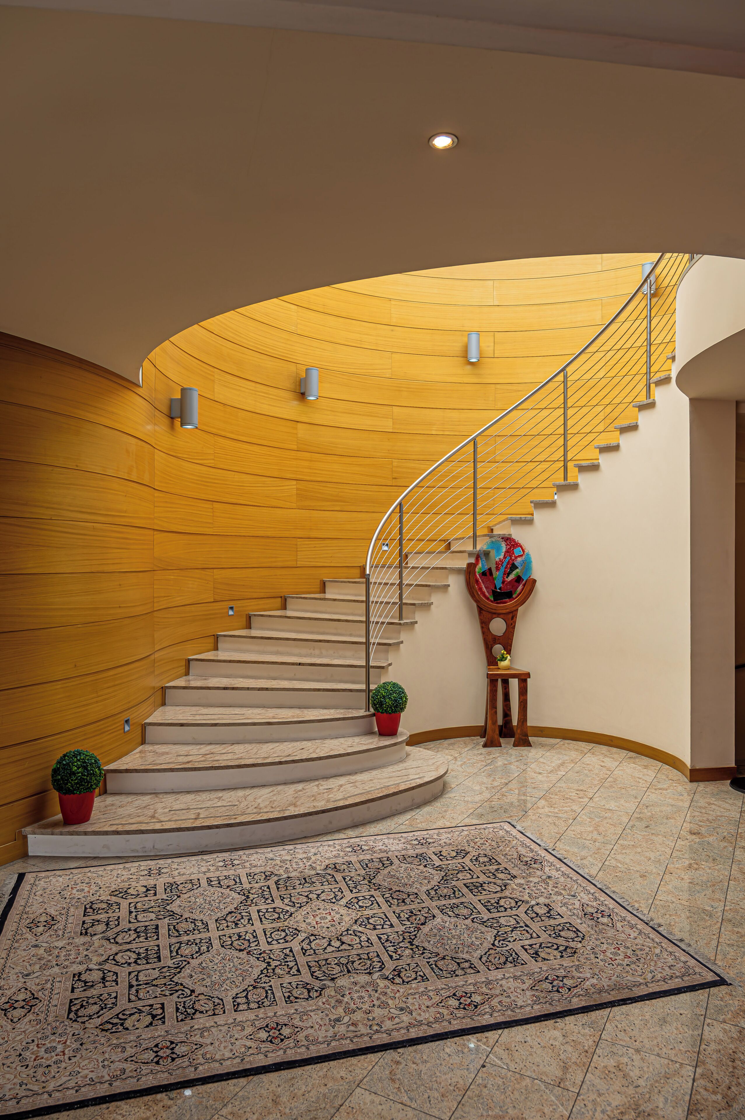 A curved staircase with a wooden wall, a metal railing, and a patterned rug on the floor.