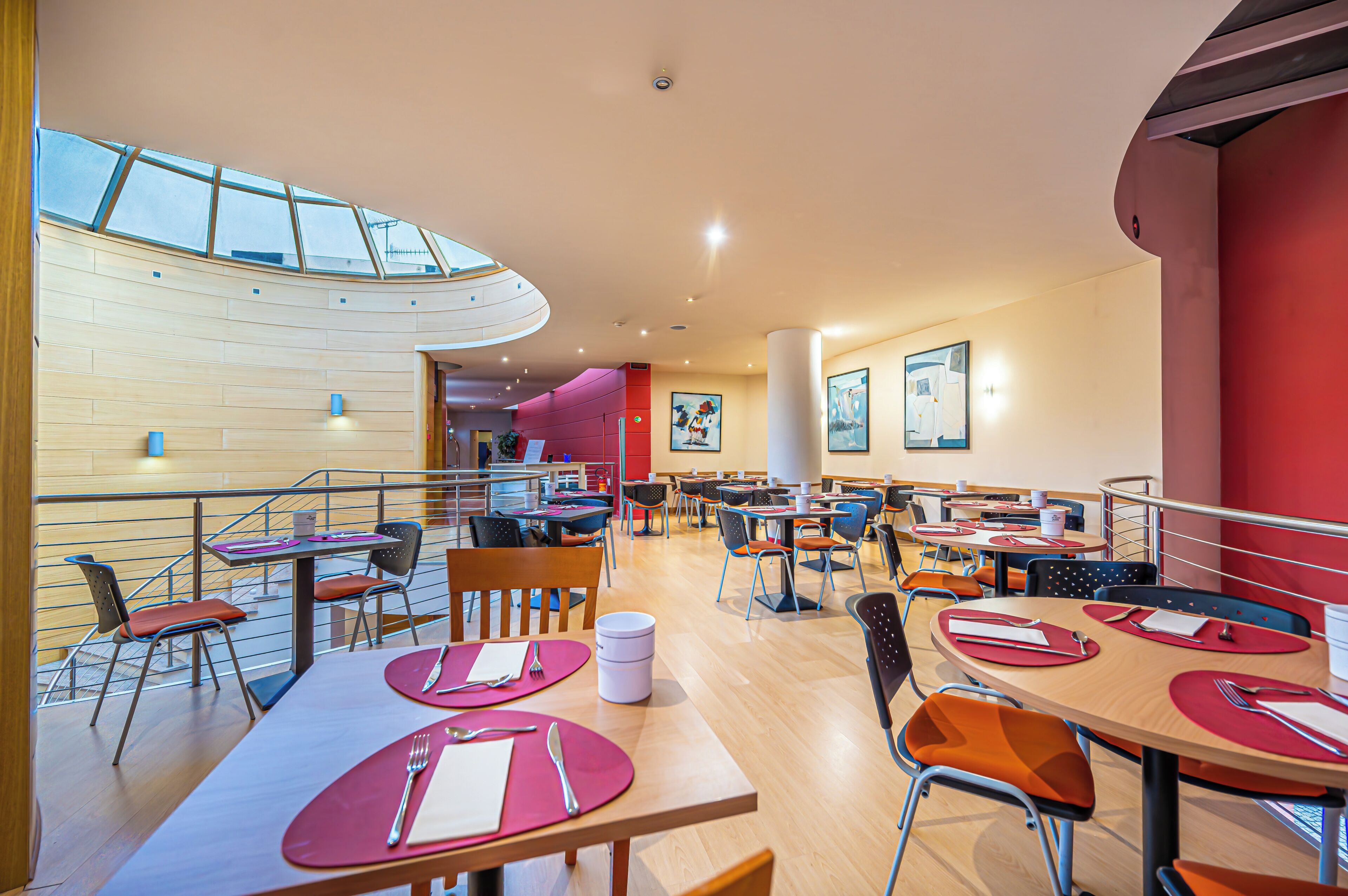Een moderne cafetaria met meerdere tafels en stoelen, een trap en een hoge plafond met dakramen.