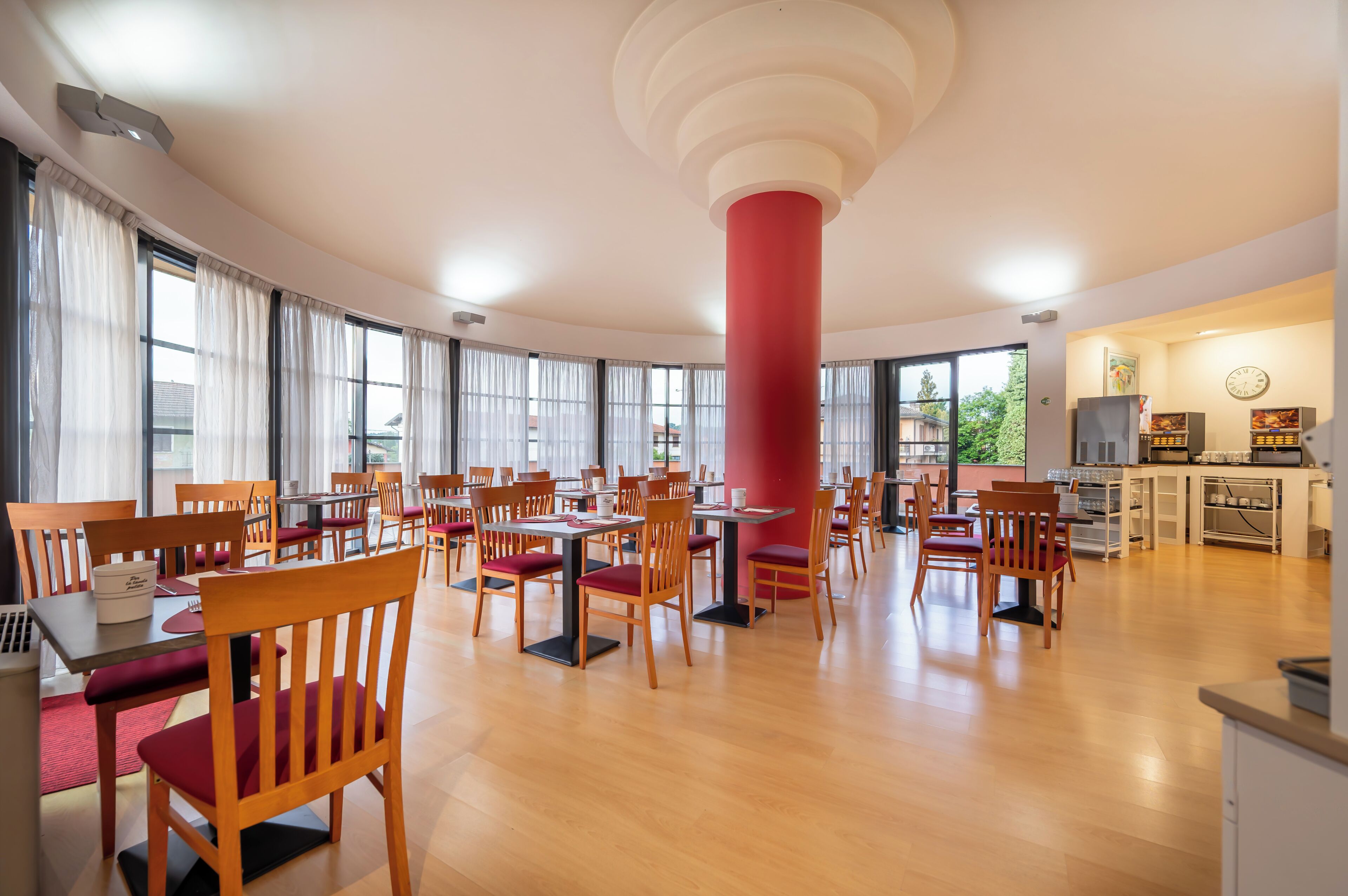 A spacious dining area with wooden chairs and tables, large windows, and a central red pillar.