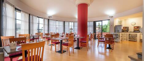 A spacious dining area with wooden chairs and tables, large windows, and a central red pillar.