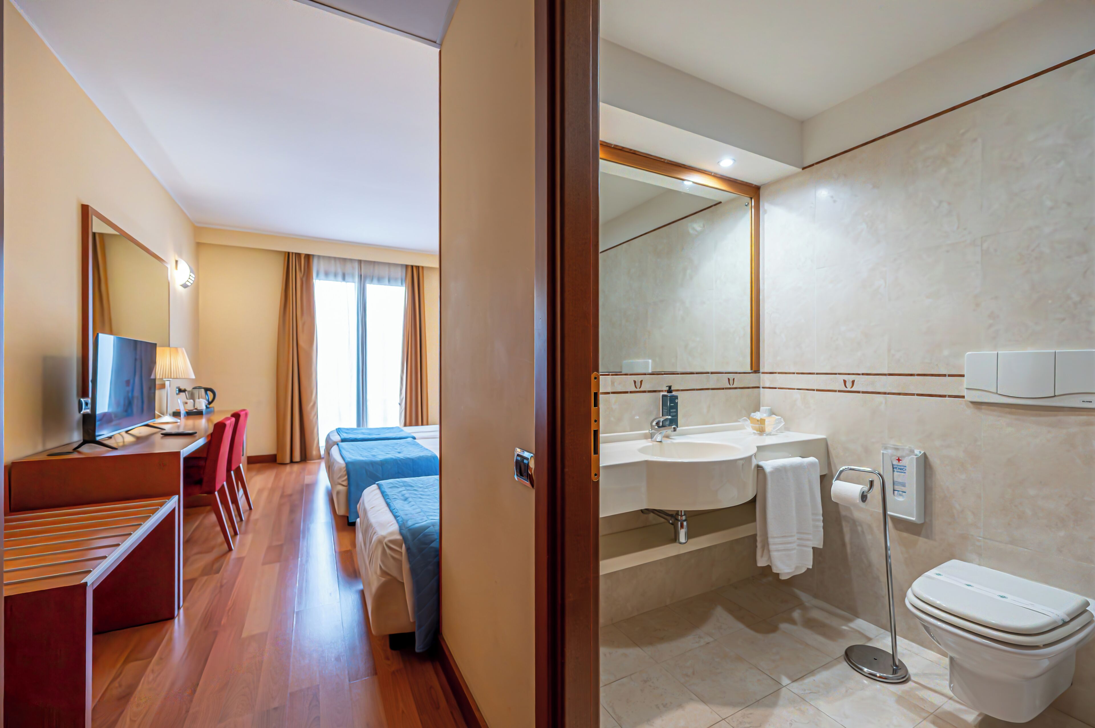 A hotel room with a desk, chairs, a bed, a bathroom with a sink, mirror, and toilet.