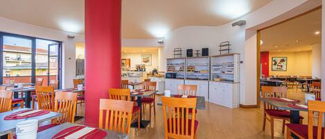 A restaurant with multiple dining tables and chairs, a central red pillar, and a kitchen area in the background.