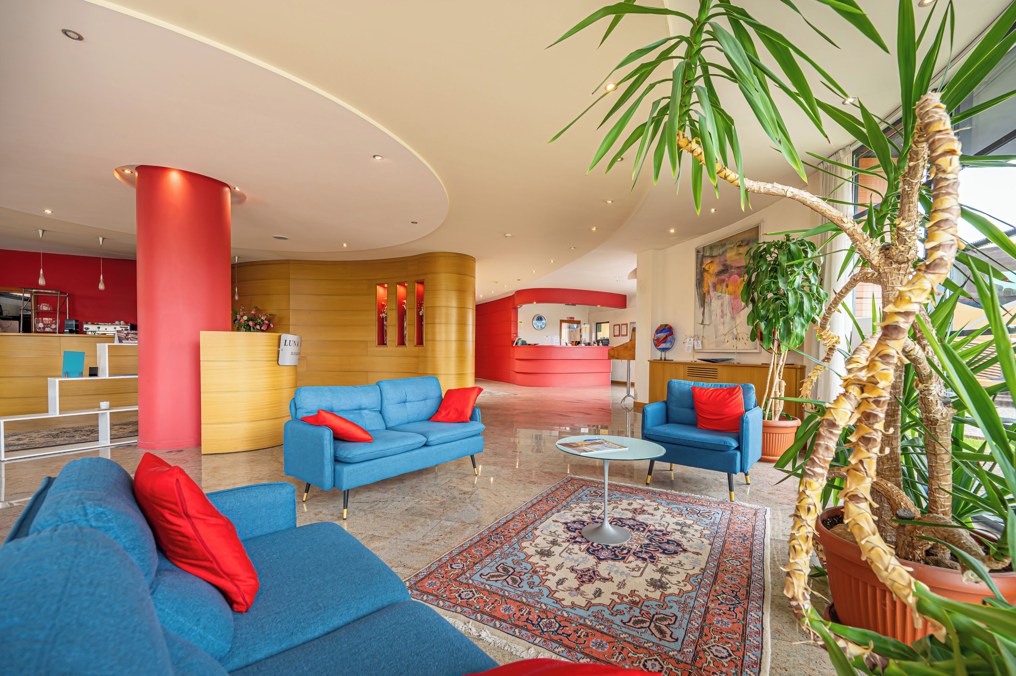 A modern lobby with blue sofas, a red reception desk, and a large potted plant.