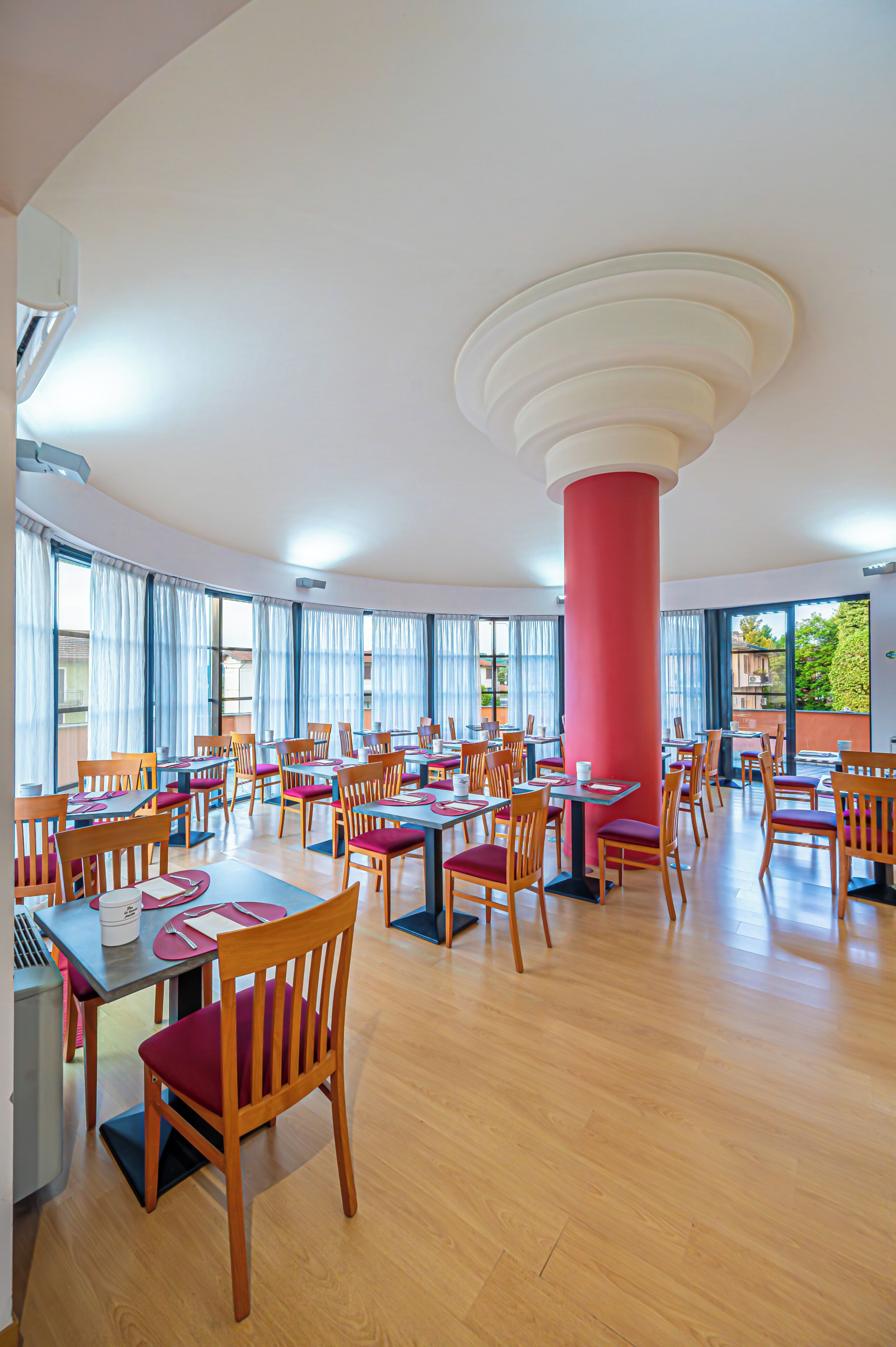 Un comedor amplio con varias mesas y sillas, ventanas grandes y un pilar rojo destacado.