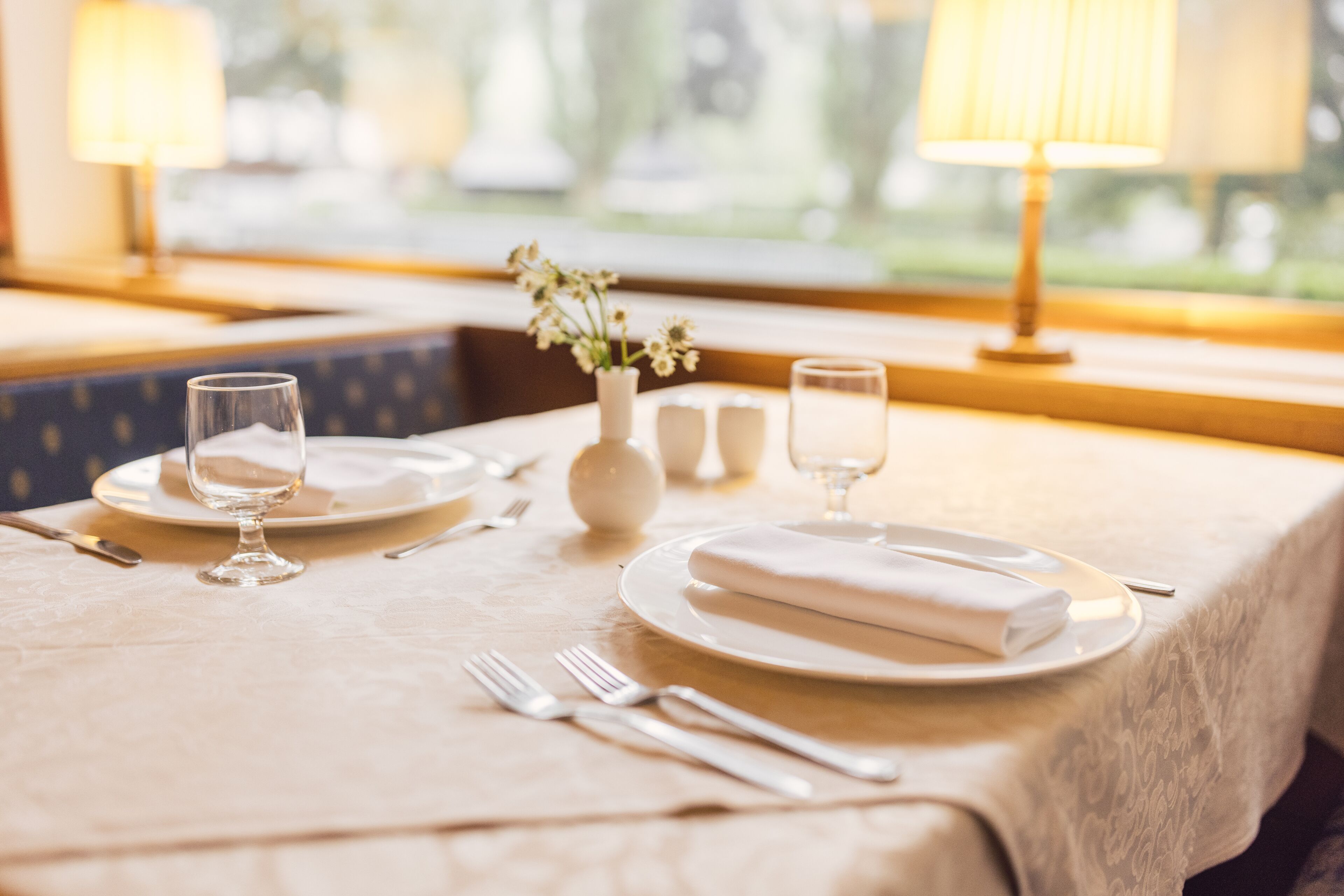Mesa de jantar posta para dois, com pratos, copos e talheres, um vaso com flores e vista pela janela.