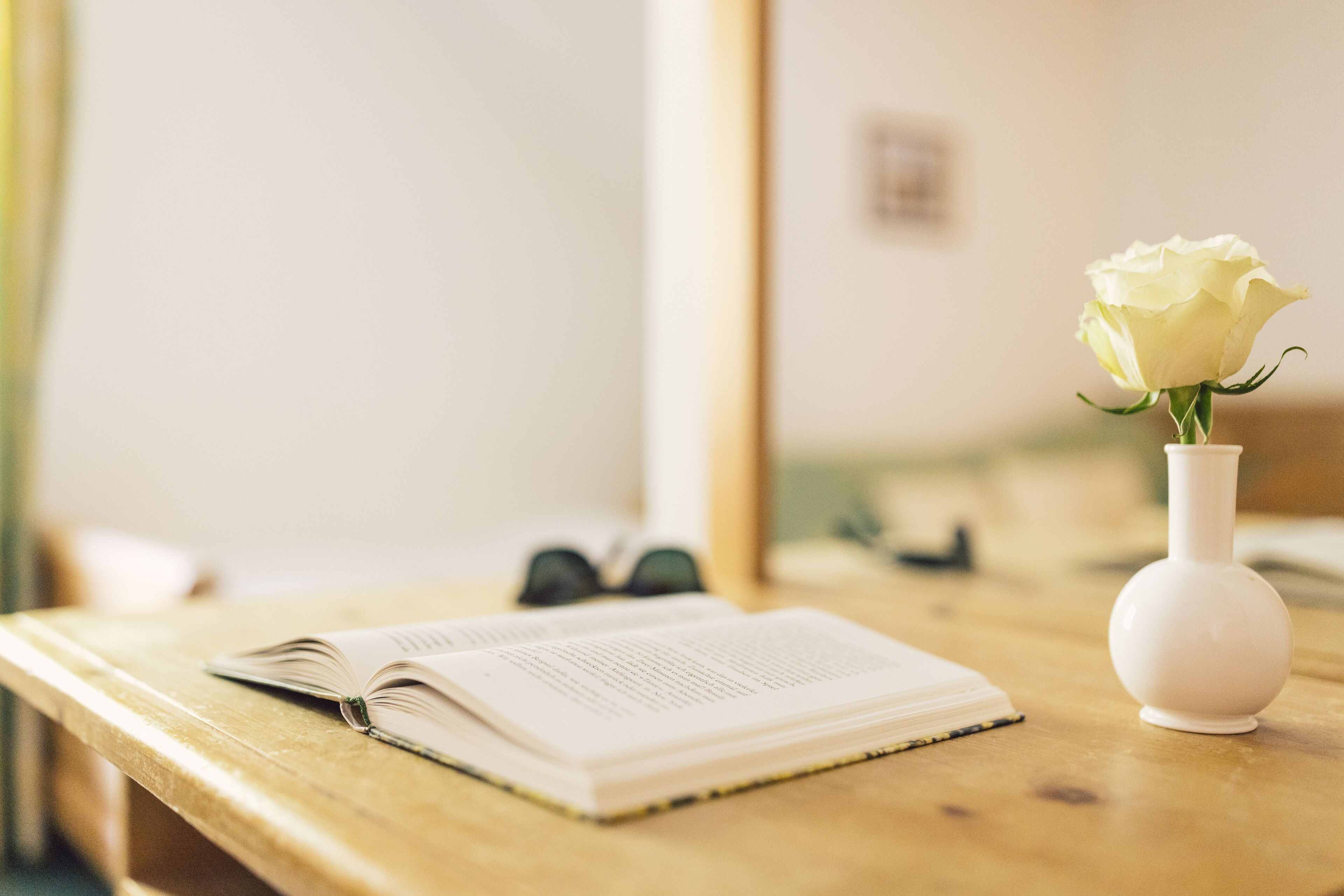 A wooden table with an open book, a white vase with a single white rose, and a pair of glasses.