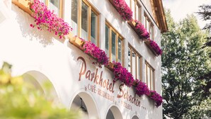 A building with arched windows and balconies with pink flowers. The sign reads "Parkhotel Schachen" and mentions a cafe, restaurant, and pizzeria.