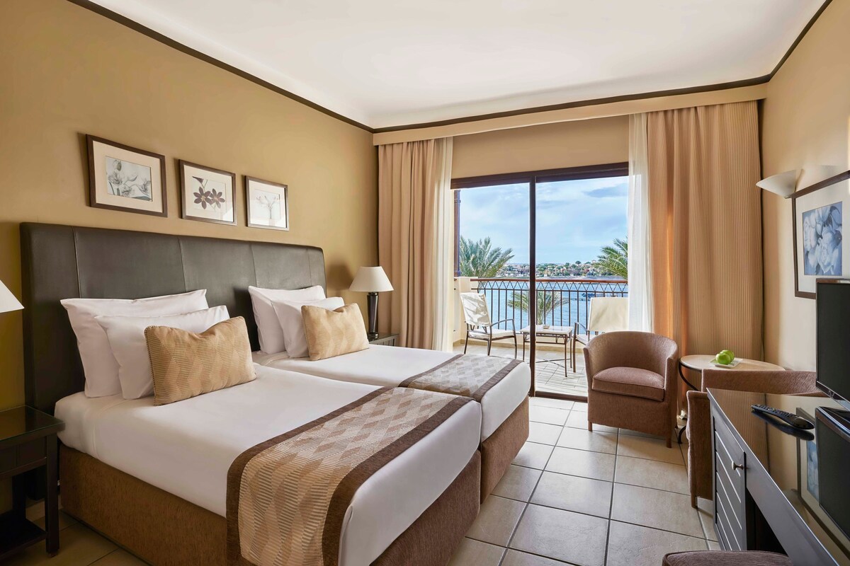 A hotel room with two beds, a balcony with a chair, a TV, and framed artwork on the wall.