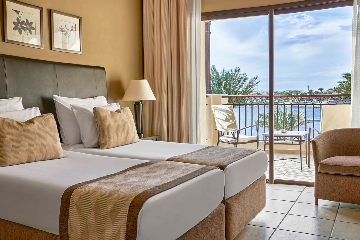 A hotel room with two beds, a balcony with a table and chairs, and a view of the sea.
