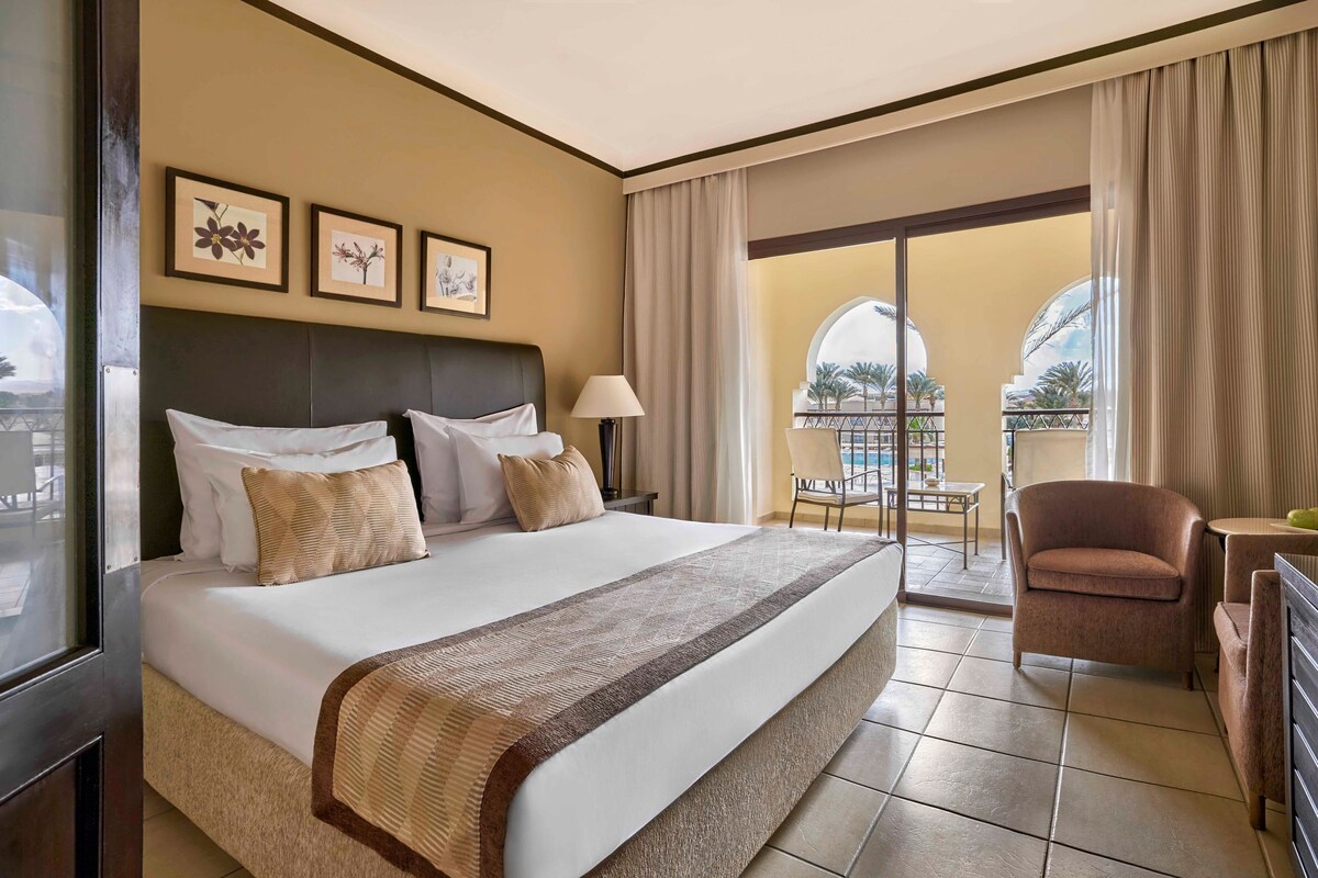 A hotel room with a large bed, a chair, a desk, and a balcony with a view.