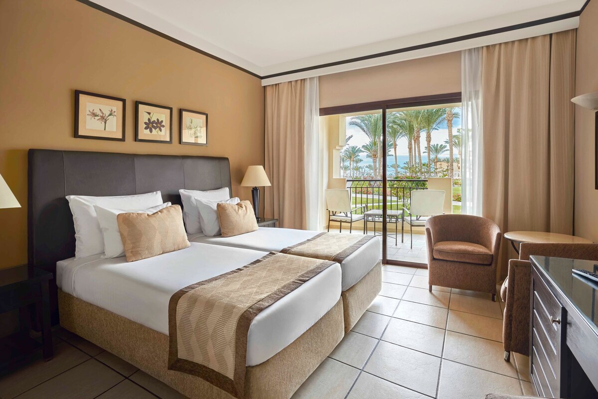 A hotel room with two beds, a desk, a chair, and a balcony with a view of palm trees and the ocean.