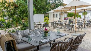 An outdoor dining area with tables and chairs, surrounded by greenery and umbrellas.