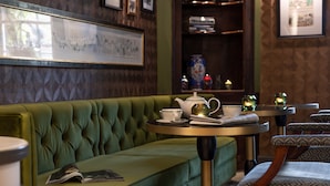 A green upholstered sofa, a round table with a teapot and cups, and a bookshelf with decorative items. - The Relais Henley (Henley-on-Thames)