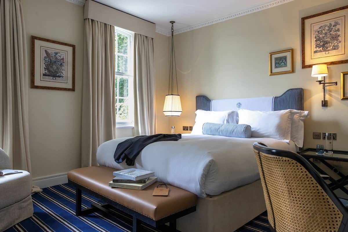 A hotel room with a large bed, a bench with books, a chair, a desk with a lamp, and framed artwork on the walls.