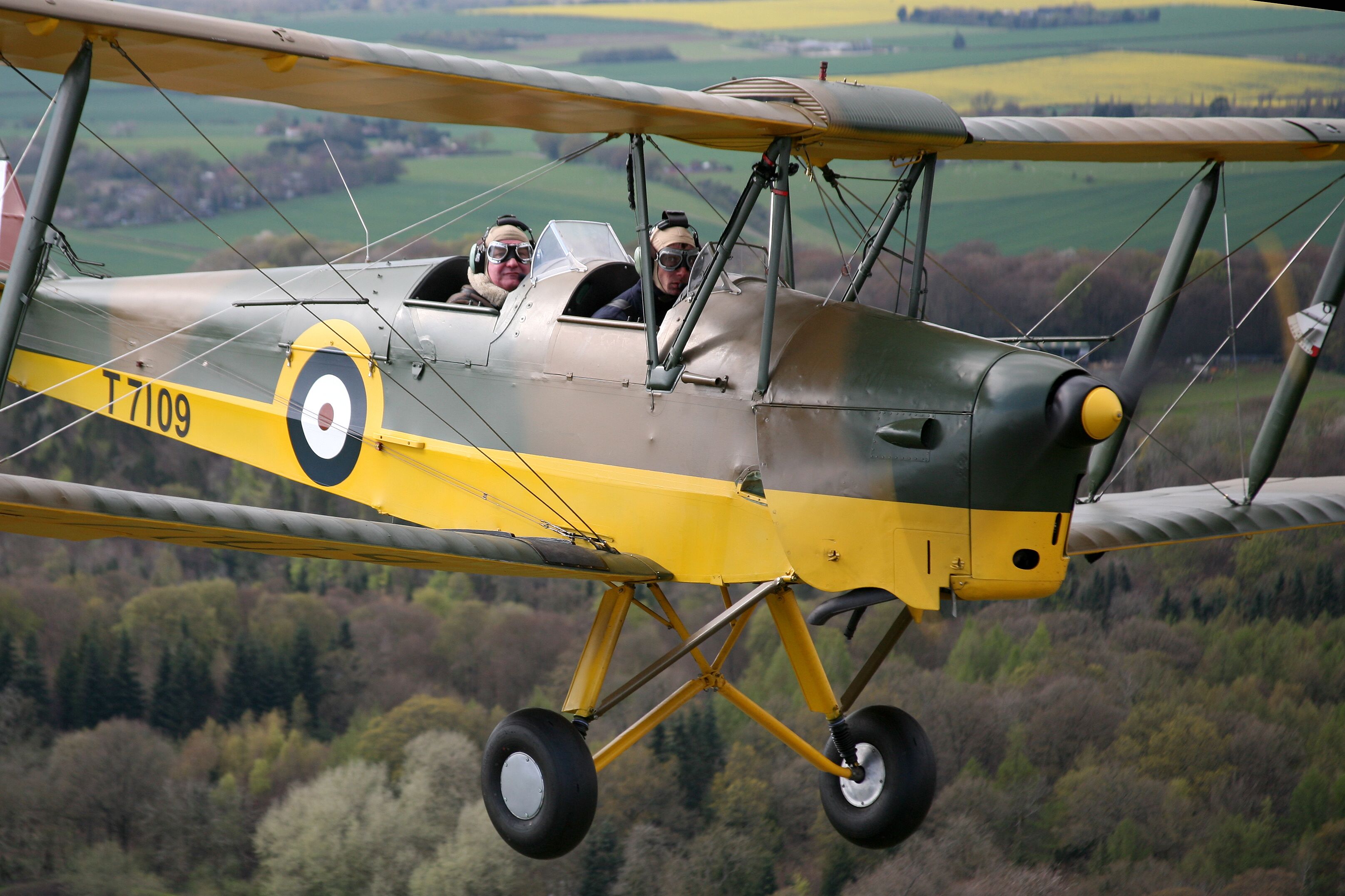 A vintage biplane with two pilots, registration T7109, flying over a rural landscape.