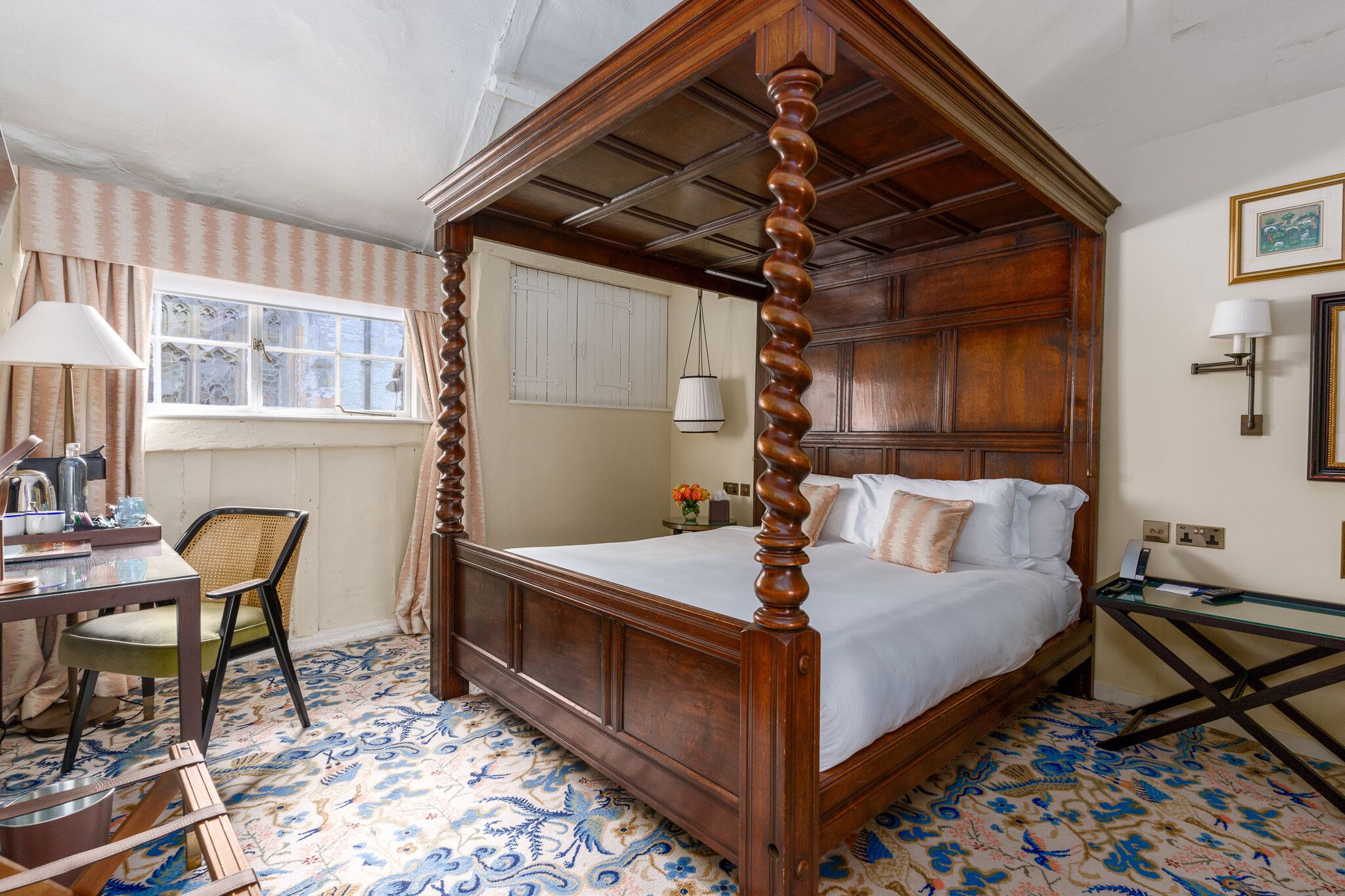 A four-poster bed with a wooden frame, a desk with a lamp, a chair, and a patterned floor.