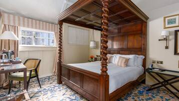 A four-poster bed with a wooden frame, a desk with a lamp, a chair, and a patterned floor.