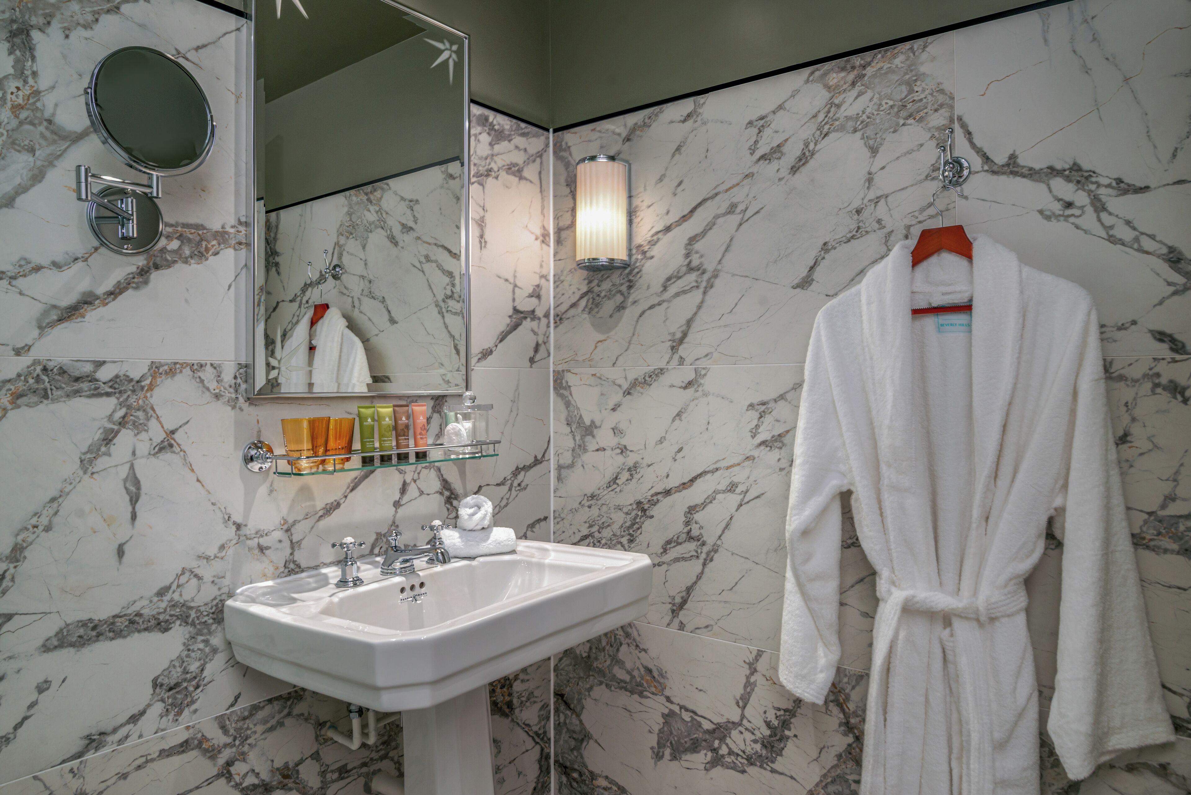 A bathroom with a marble wall, a sink, a mirror, a robe hanging, and a towel rack.