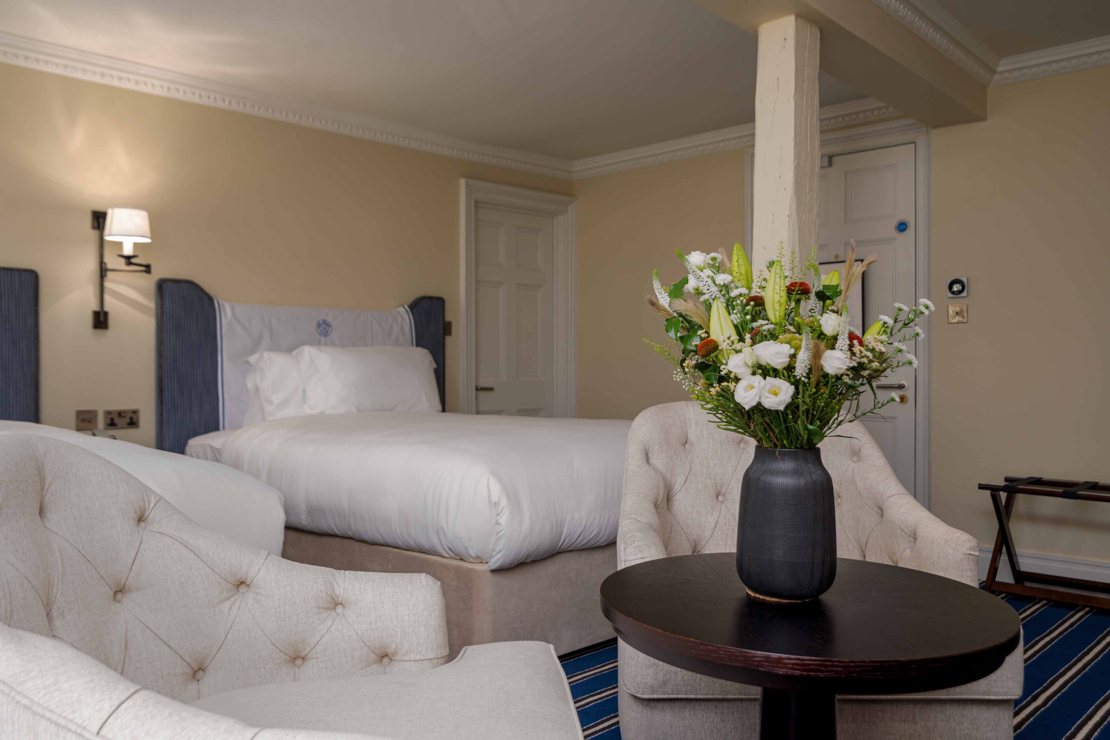 Habitación de hotel con dos camas, un área de descanso con una silla y una mesa, y un jarrón con flores sobre la mesa.