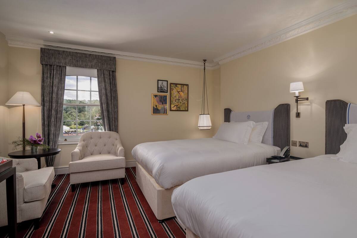 A hotel room with two beds, a chair, a table, a lamp, and a window with curtains.