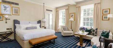 Habitación de hotel con una cama grande, un banco, un sofá, una mesita y una silla. En las paredes hay cuadros enmarcados y una ventana con cortinas.
