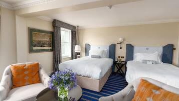 Habitación de hotel con dos camas, una zona de estar con una silla y una mesa, y un cuadro en la pared.