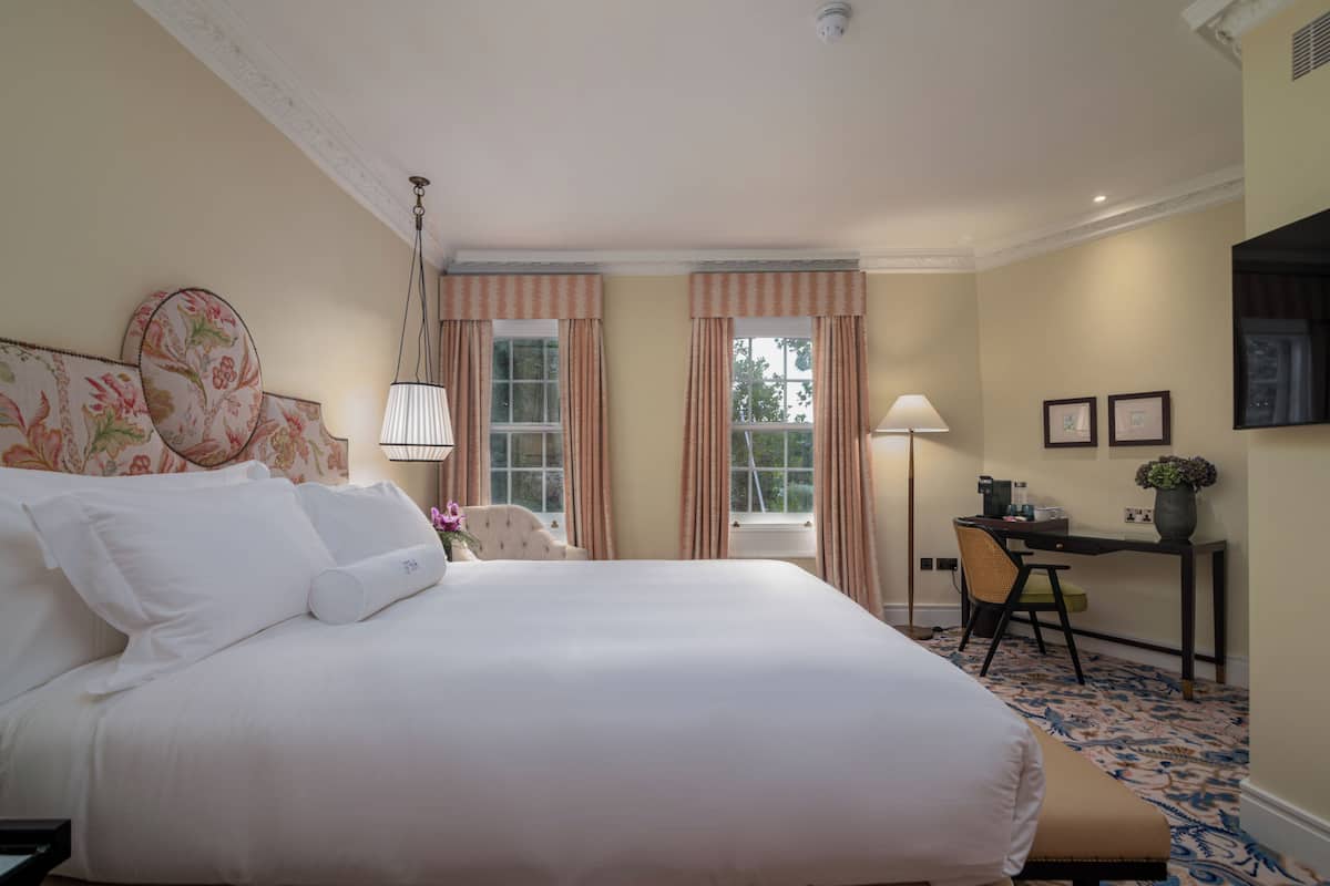 A hotel room with a large bed, a desk with a chair, and two windows with curtains.