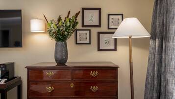 A wooden dresser with drawers, a vase with flowers, framed botanical prints on the wall, a standing lamp, and a television on a stand.