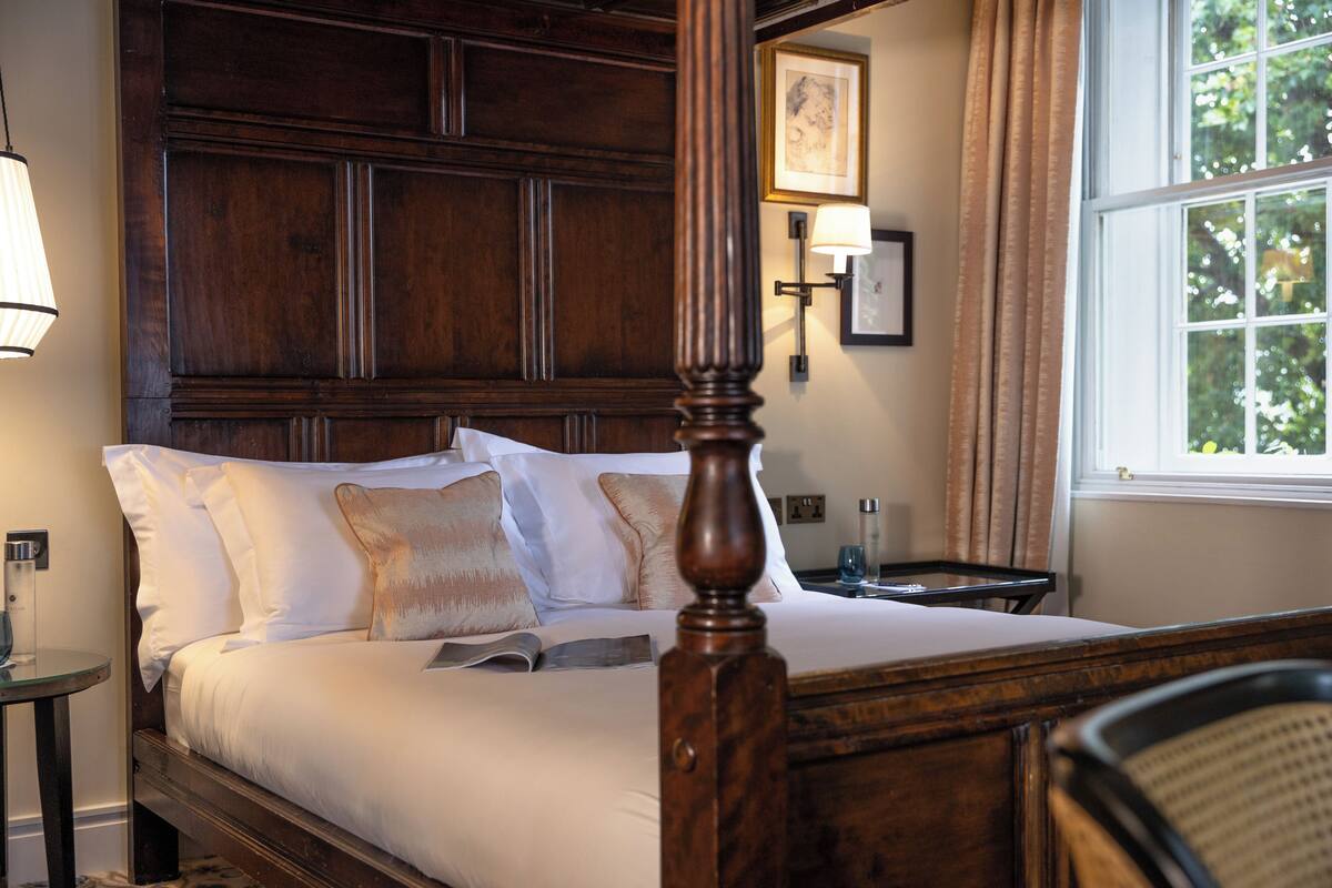 A neatly made bed with white linens and multiple pillows, a wooden headboard, a bedside table with a lamp, and a window with curtains.