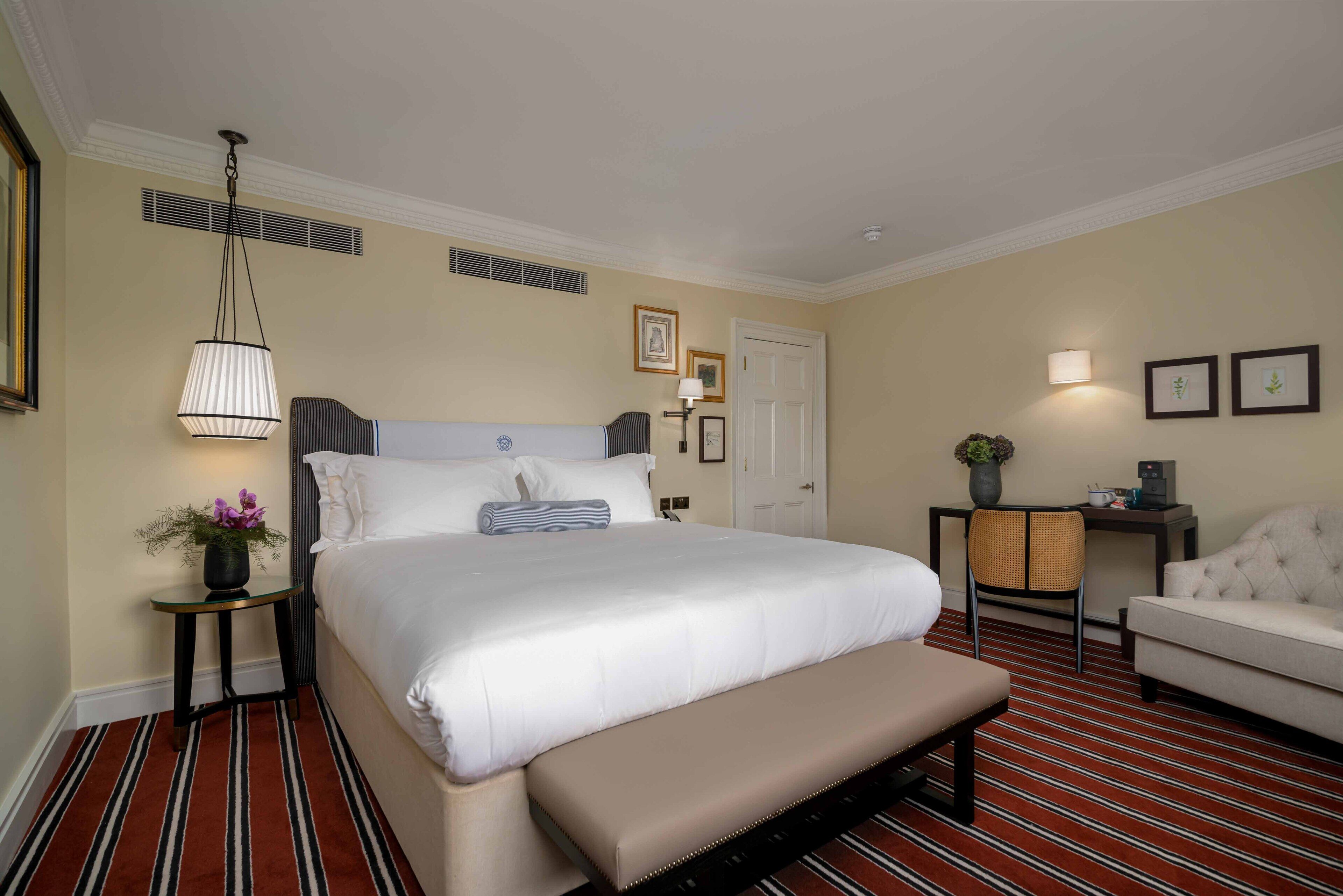 Habitación de hotel con una cama grande, un banco al pie de la cama, una mesita de noche con lámpara, una silla y un sofá.
