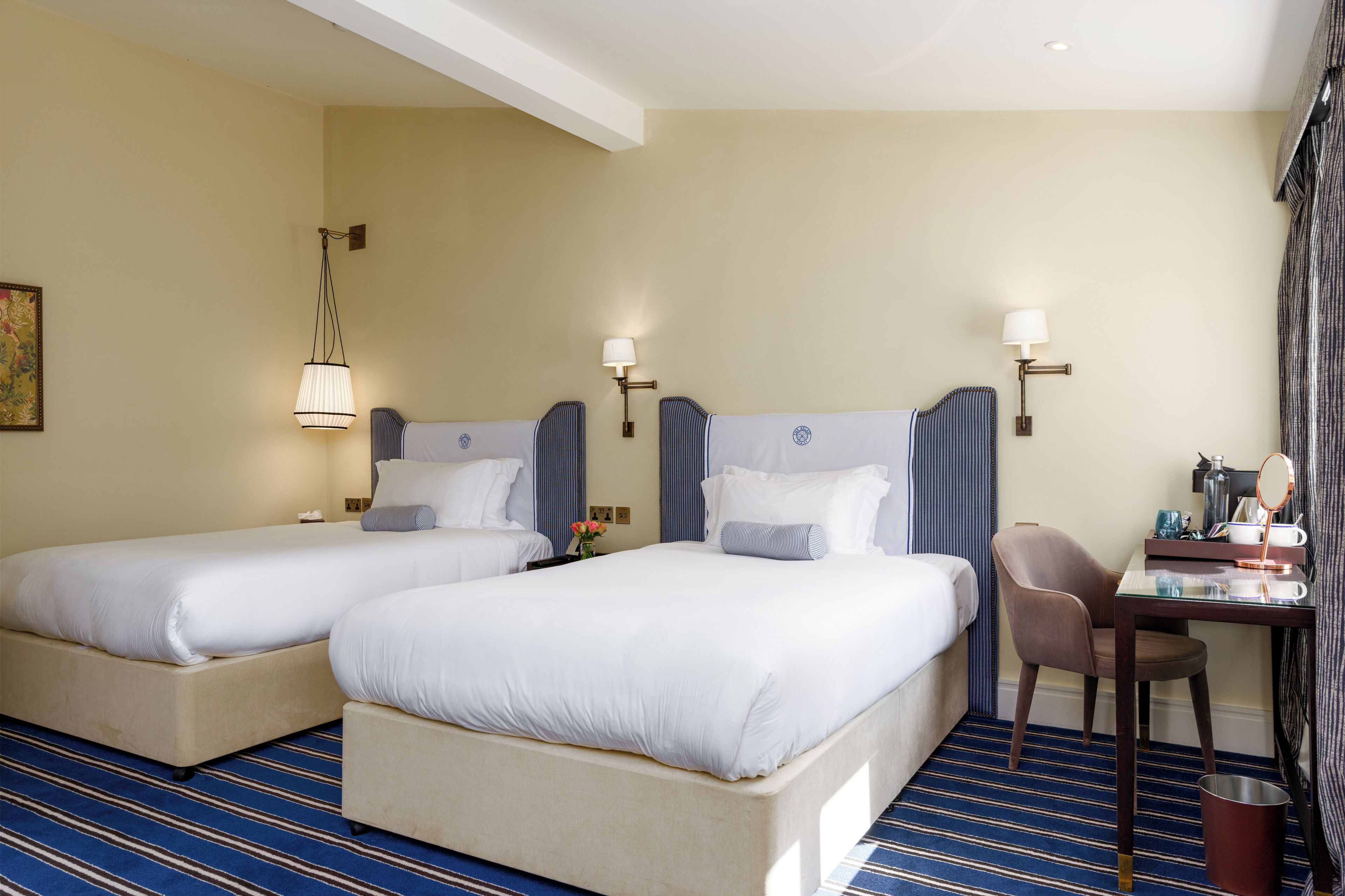 Habitación de hotel con dos camas, un escritorio con silla, y una mesita de noche con espejo y artículos de aseo.