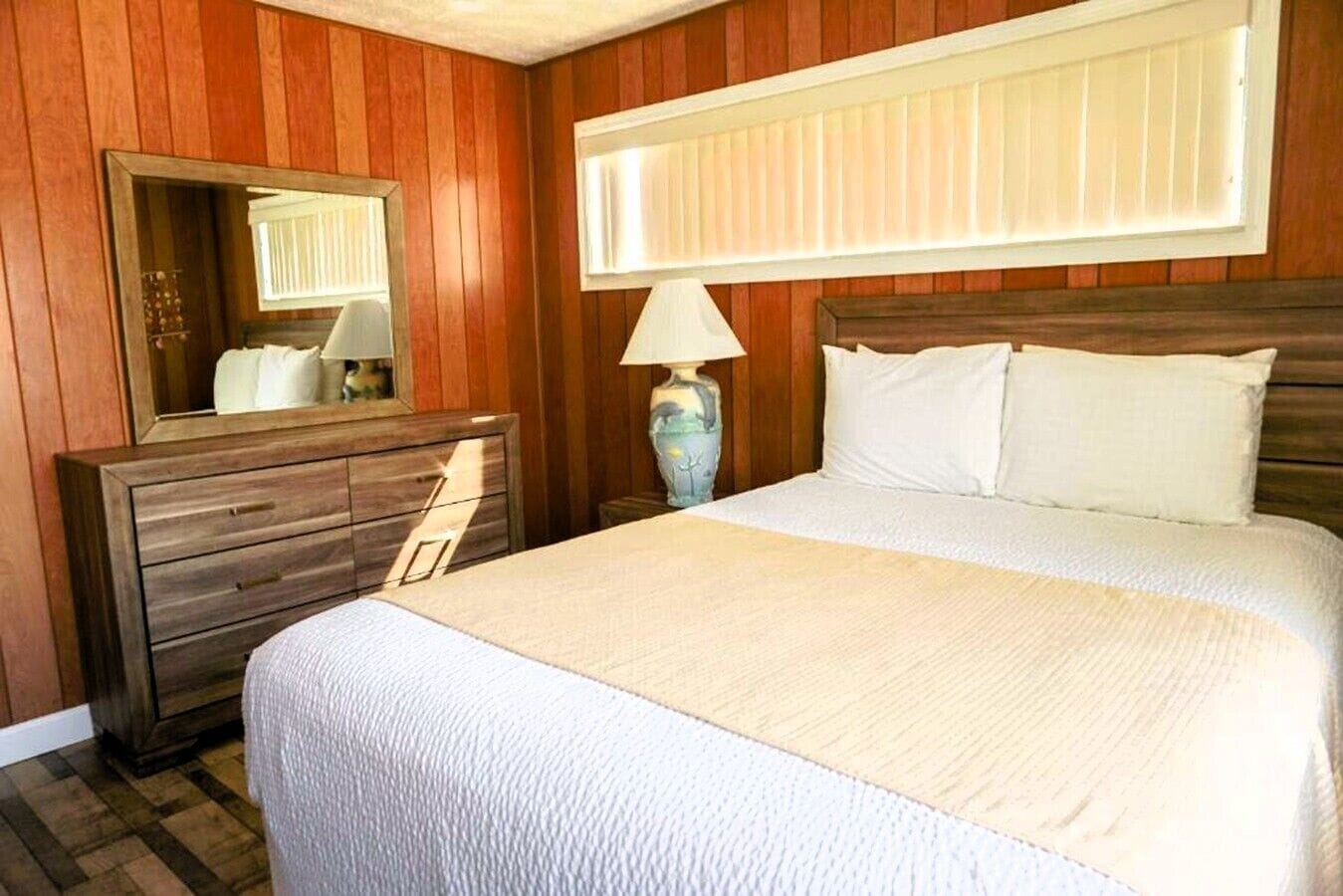 Une chambre d’hôtel avec un mur lambrissé en bois, un grand lit avec une literie blanche, une commode avec un miroir et une fenêtre avec des stores.