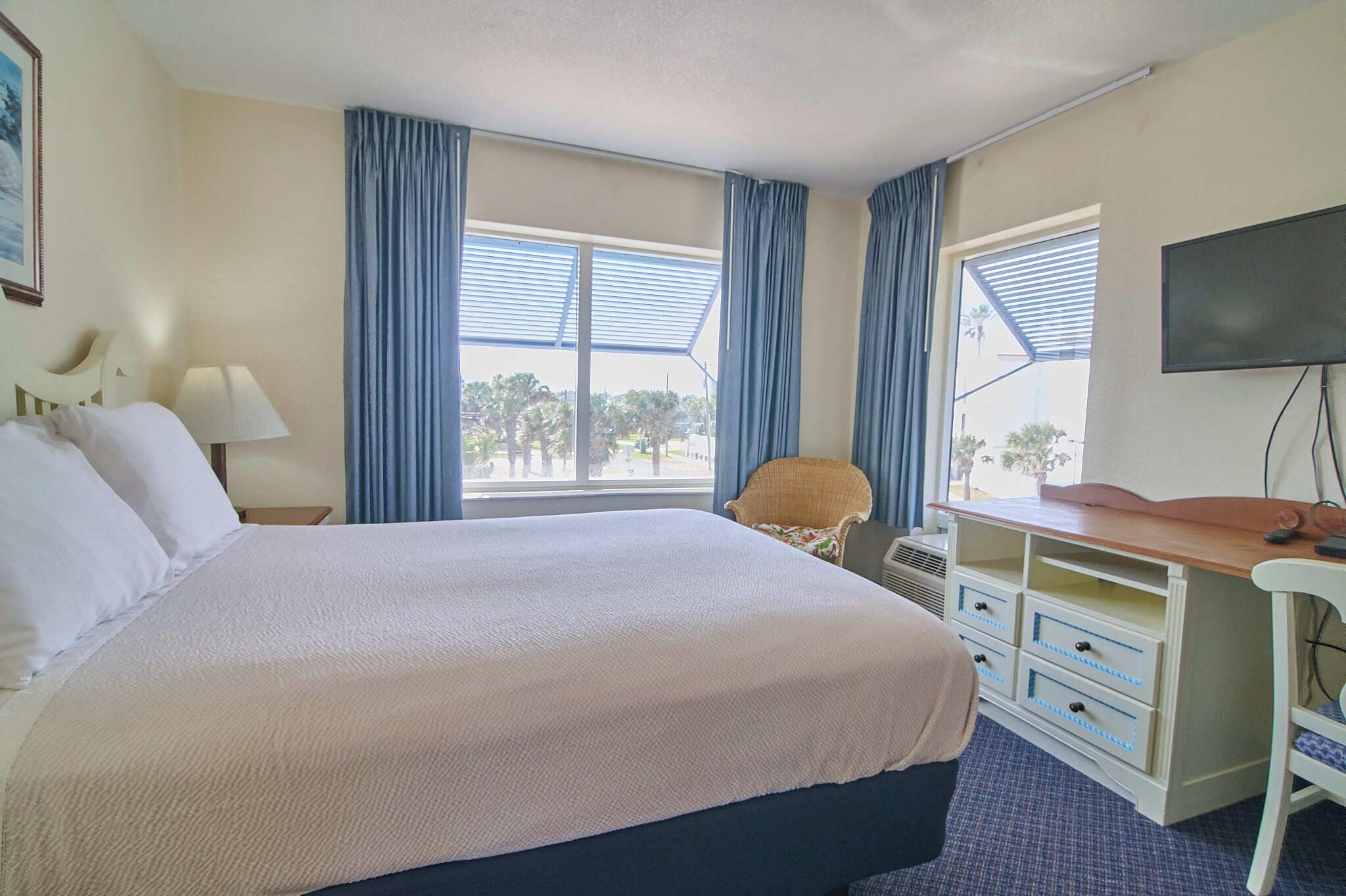 A hotel room with a large bed, a desk with drawers, a chair, a TV, and a window with curtains.