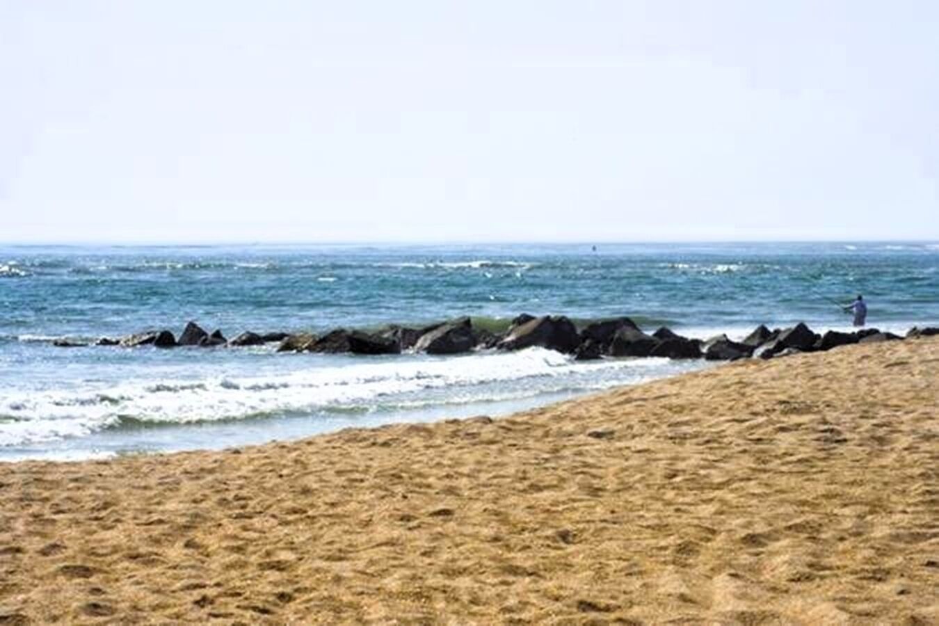 A sandy beach with a rocky shoreline and a person fishing in the water.