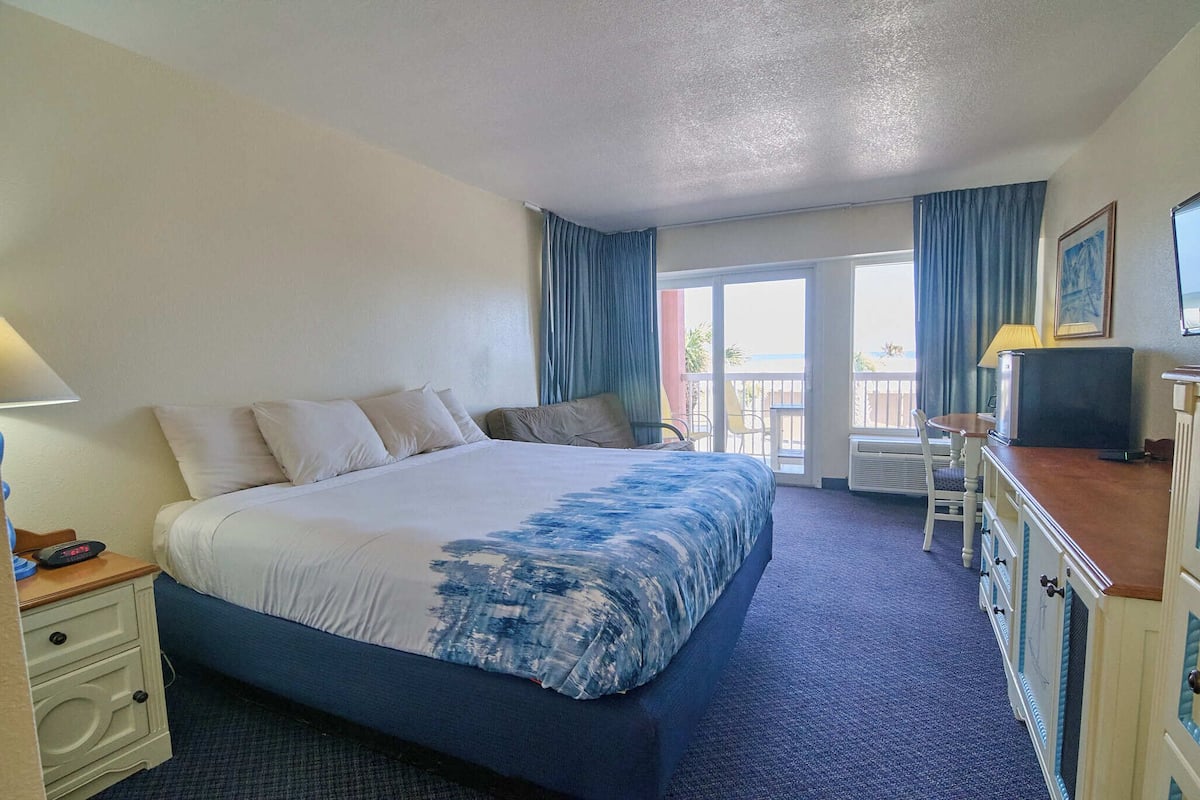 A hotel room with a large bed, a desk with a lamp, a television, and a balcony with a view.