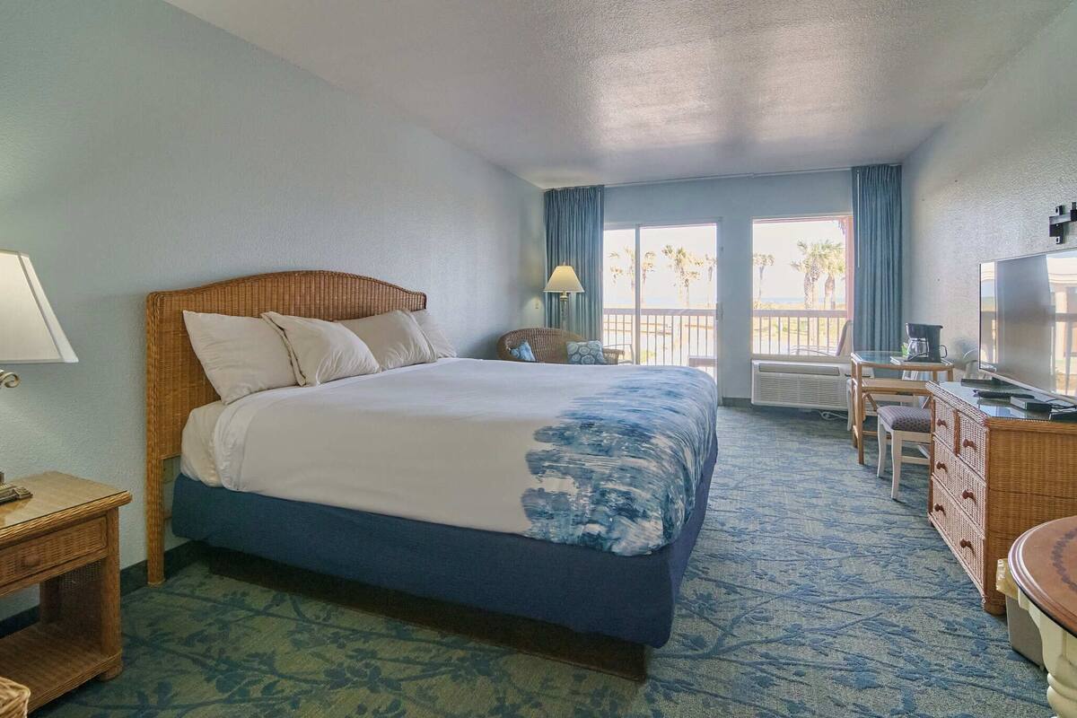 A hotel room with a bed, a desk, a chair, a lamp, and a view of the outdoors.