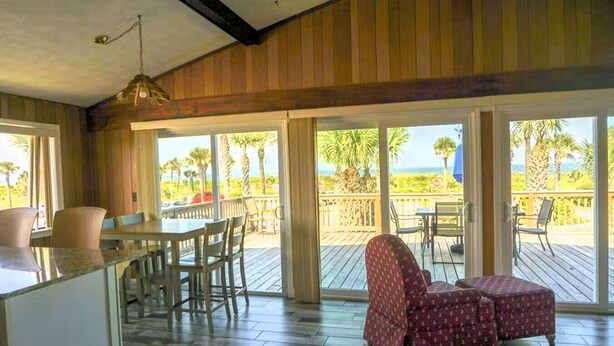 A room with a wooden ceiling, large windows, a dining area with chairs, a table, and a patterned sofa.