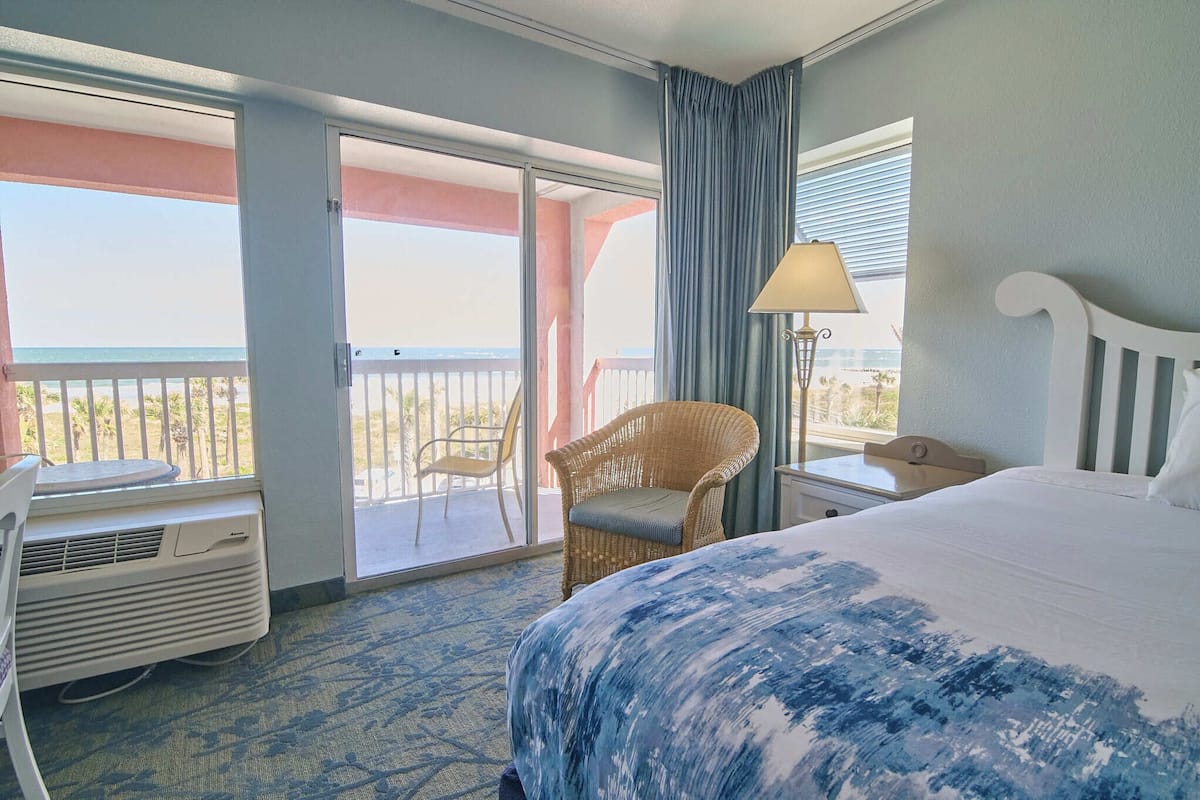 A hotel room with a large bed, a wicker chair, a table, and a view of the beach through a sliding glass door.