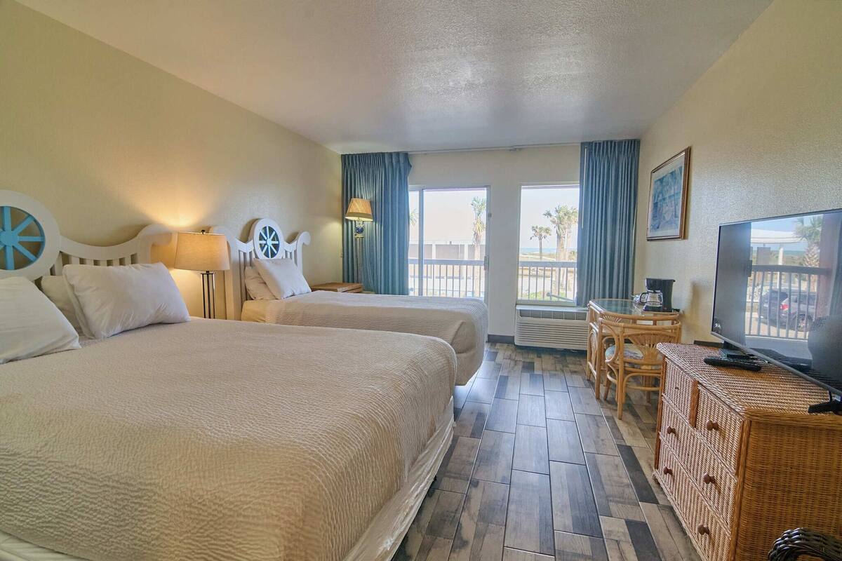 A hotel room with two beds, a television, a desk, and a view of the beach.