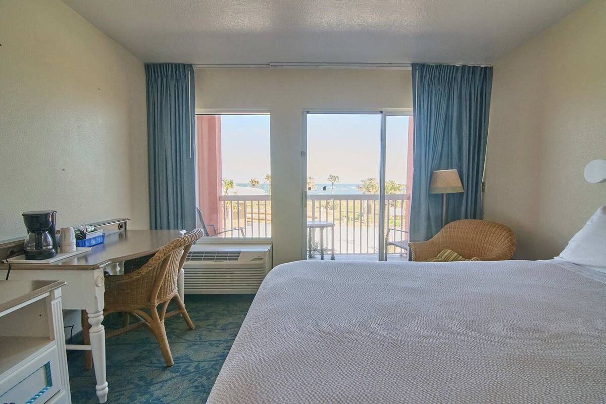 A hotel room with a bed, a desk with a chair, a lamp, and a view of the beach through the balcony doors.