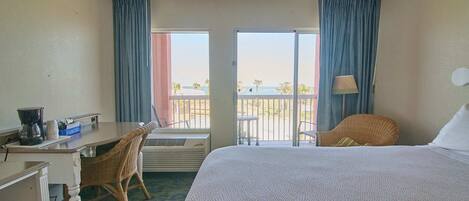 Habitación de hotel con cama, escritorio con silla, lámpara y vista a la playa a través de las puertas del balcón.
