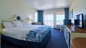 A hotel room with a bed, a desk with a television, a chair, and a view of the ocean.