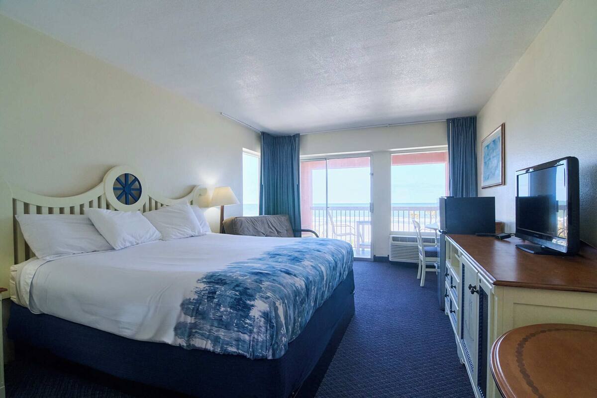 A hotel room with a bed, a desk with a television, a chair, and a view of the ocean.