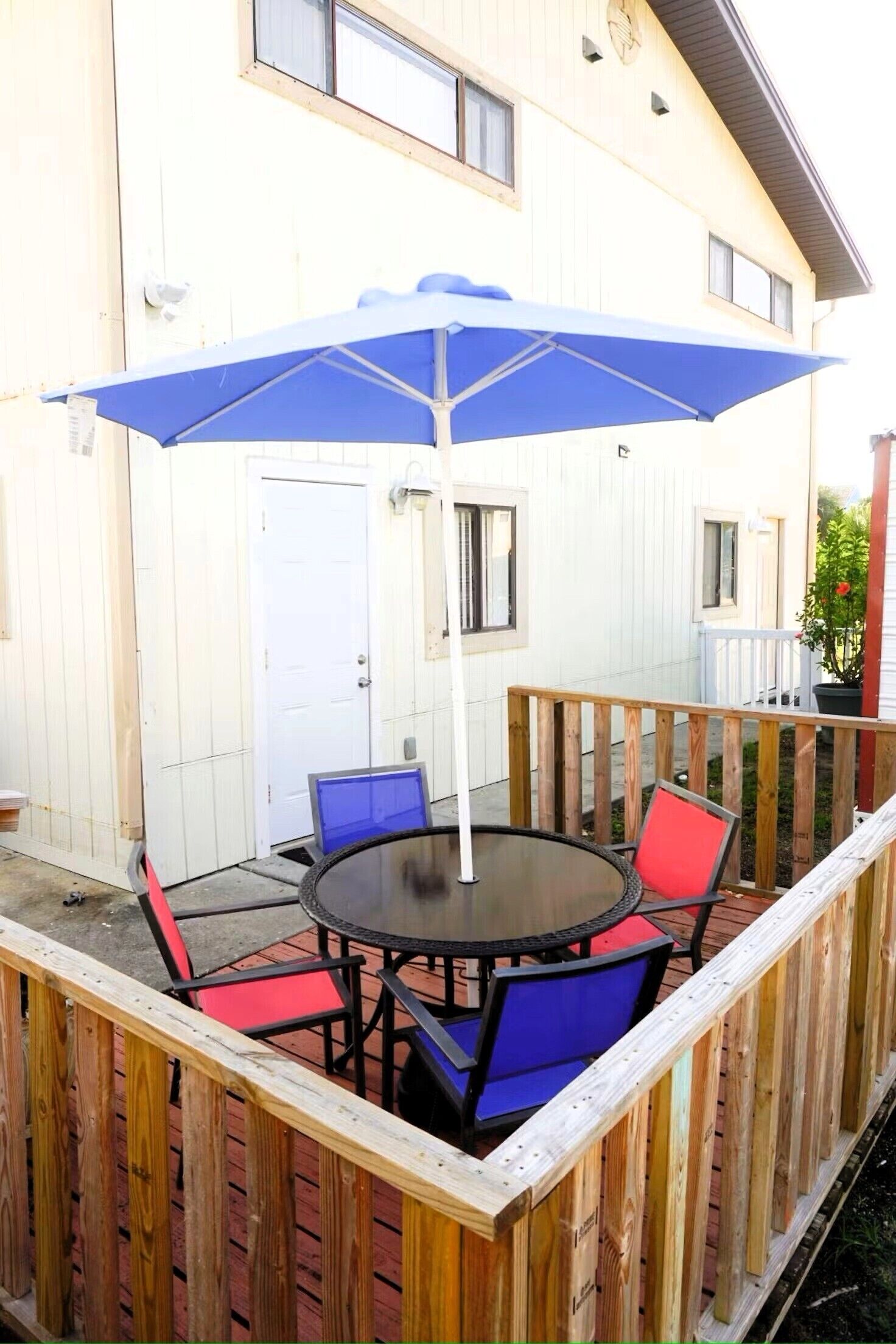 A wooden deck with a round table and chairs, a blue umbrella, and a white door.