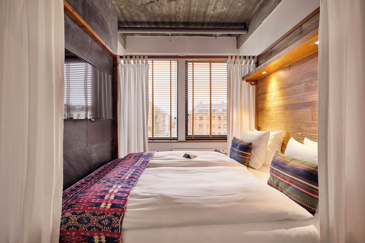 Ein modernes Hotelzimmer mit einem großen Bett, einem Holzkopfteil und Blick auf die Stadt durch raumhohe Fenster.